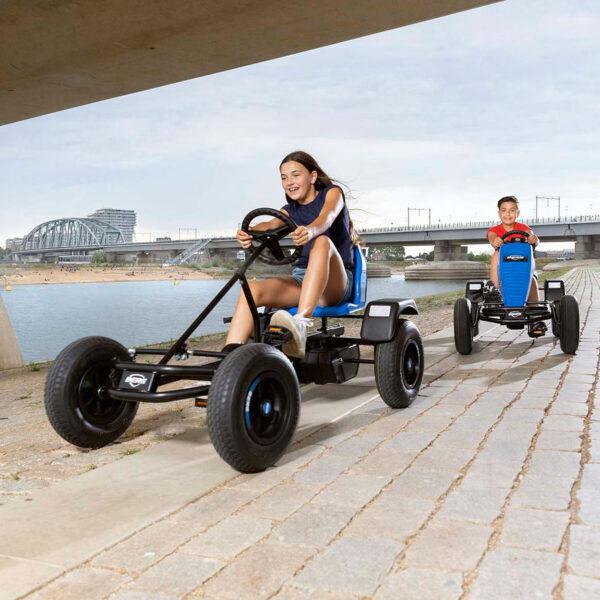 Two children race on BERG XL Basics go-karts under a bridge by the water