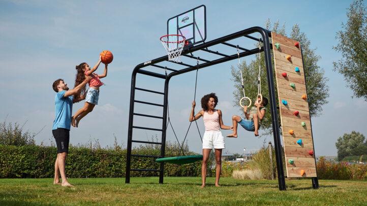 Niños jugando en un parques infantiles BERG PlayBase con aro de baloncesto, columpio y pared de escalada en el jardín