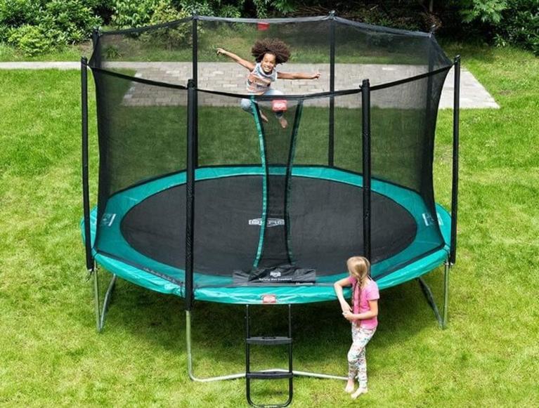 Kind springt auf einem runden BERG Trampolin mit Sicherheitsnetz, während ein anderes Kind im Garten wartet