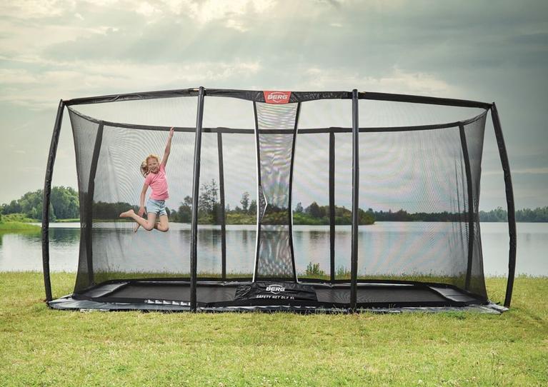 Enfant sautant sur un trampoline rectangulaire BERG avec filet dans un cadre verdoyant près de l’eau