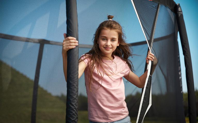 Lächelndes Mädchen öffnet das Sicherheitsnetz eines BERG Trampolins in einem sonnigen Garten