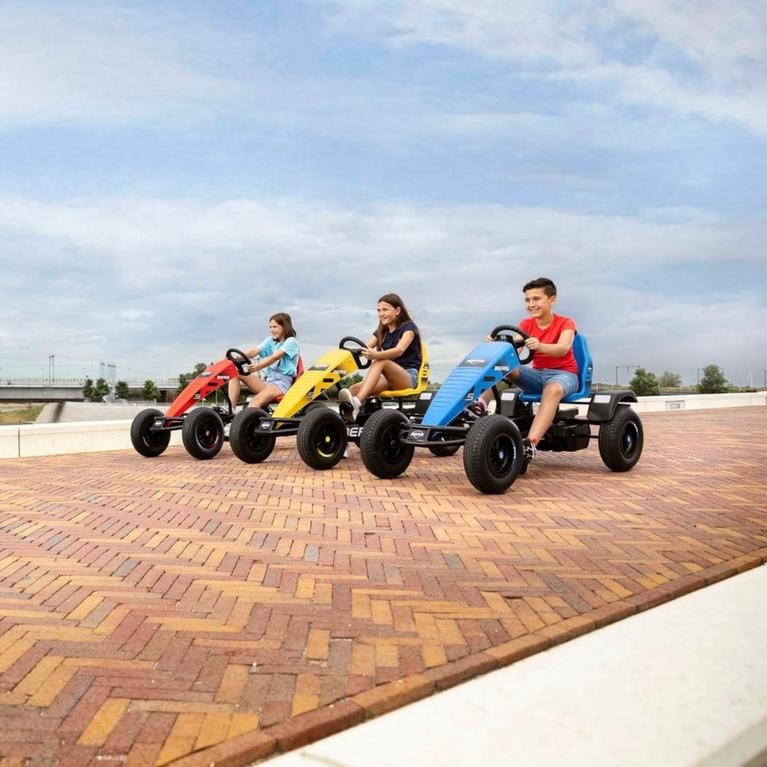 Three children racing on electric BERG pedal go-karts over a brick-paved square