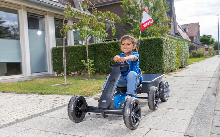 Niño feliz conduciendo un kart a pedales BERG Reppy Roadster con remolque y bandera en la acera