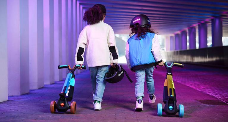 Two children walking with their BERG Nexo scooters with illuminated wheels in a colorful tunnel