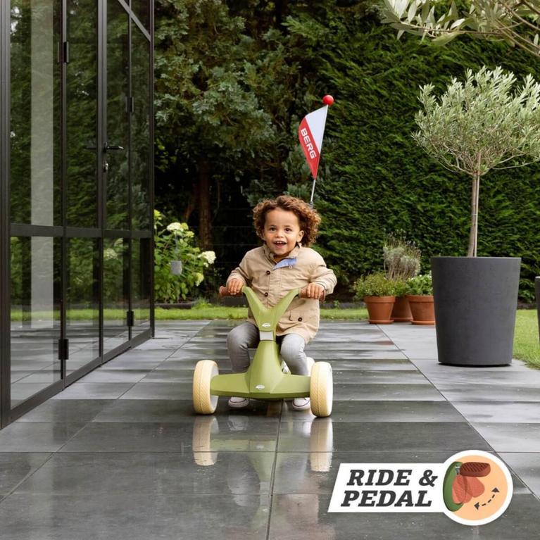 Enfant souriant en roulant sur une voiture à pousser BERG GO verte avec drapeau dans le jardin