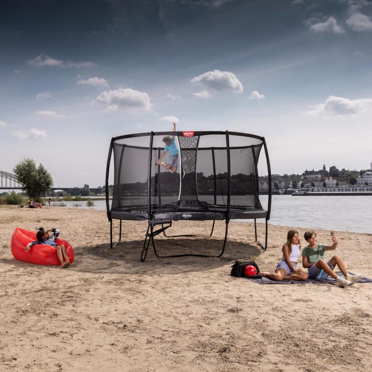 Niño saltando en un trampolín BERG Elite con red de seguridad en la playa mientras la gente se relaja