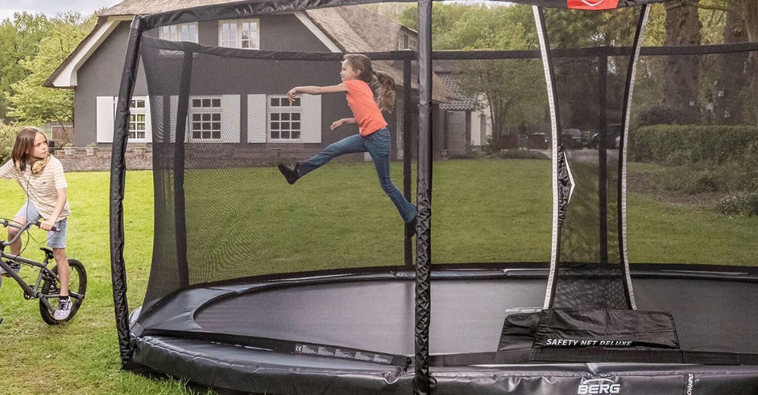 Banner mit Kindern, die auf einem BERG Champion InGround Trampolin mit Sicherheitsnetz im Garten spielen