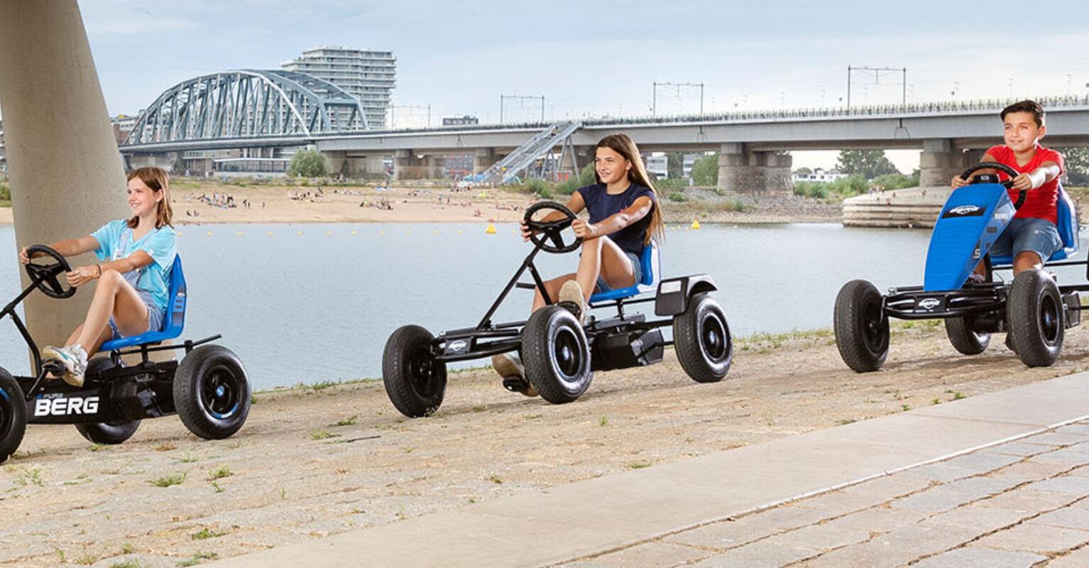 Three children drive BERG XL go-karts along a river under a bridge