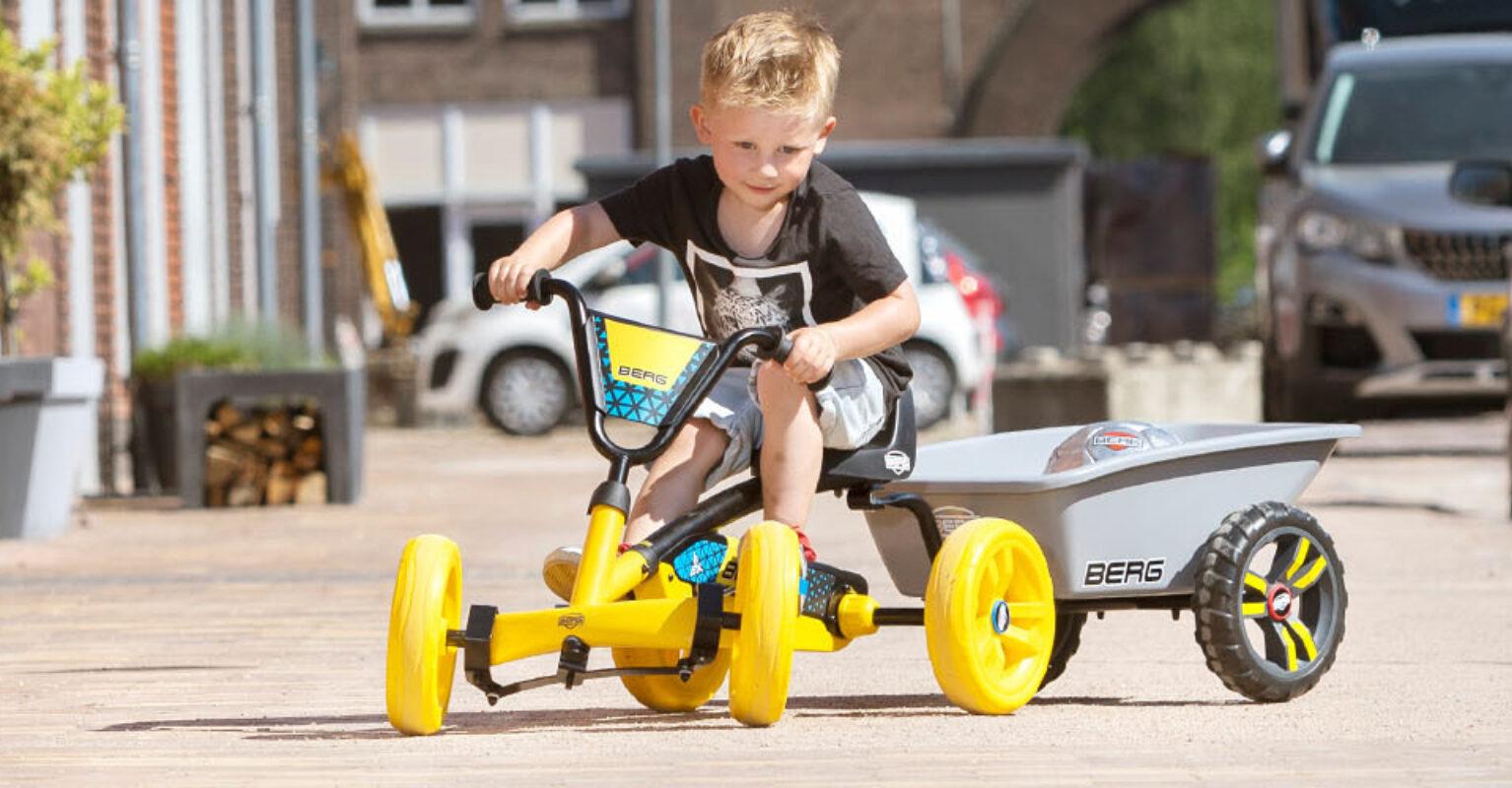 Child drives a yellow BERG Buzzy go-kart with a trailer in a residential area