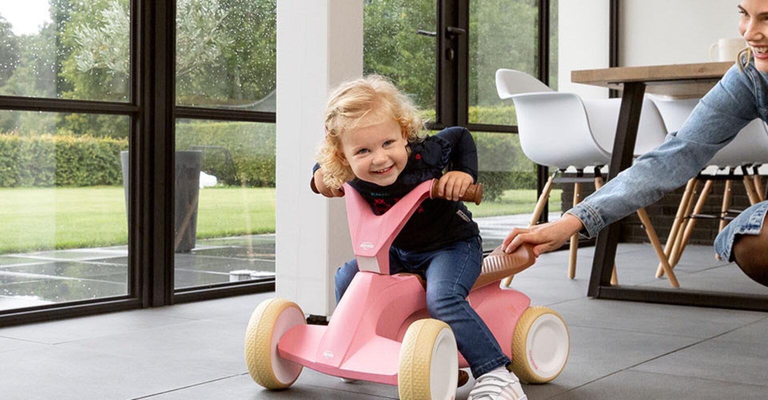 Child plays indoors with a pink BERG Go2 ride-on car while mom pushes