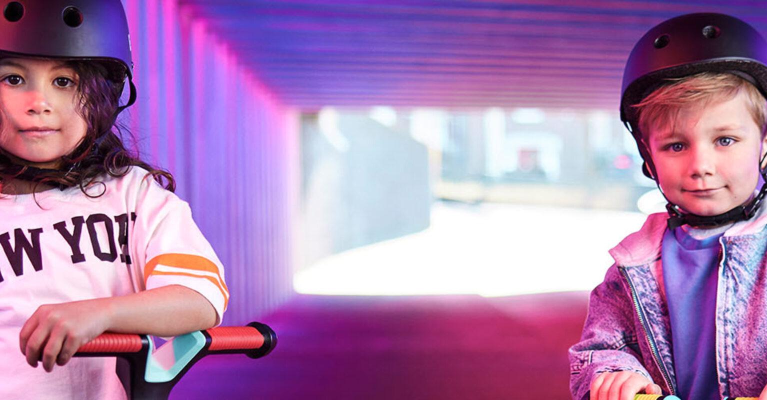 Two children ride BERG Nexo scooters under a colorfully lit underpass