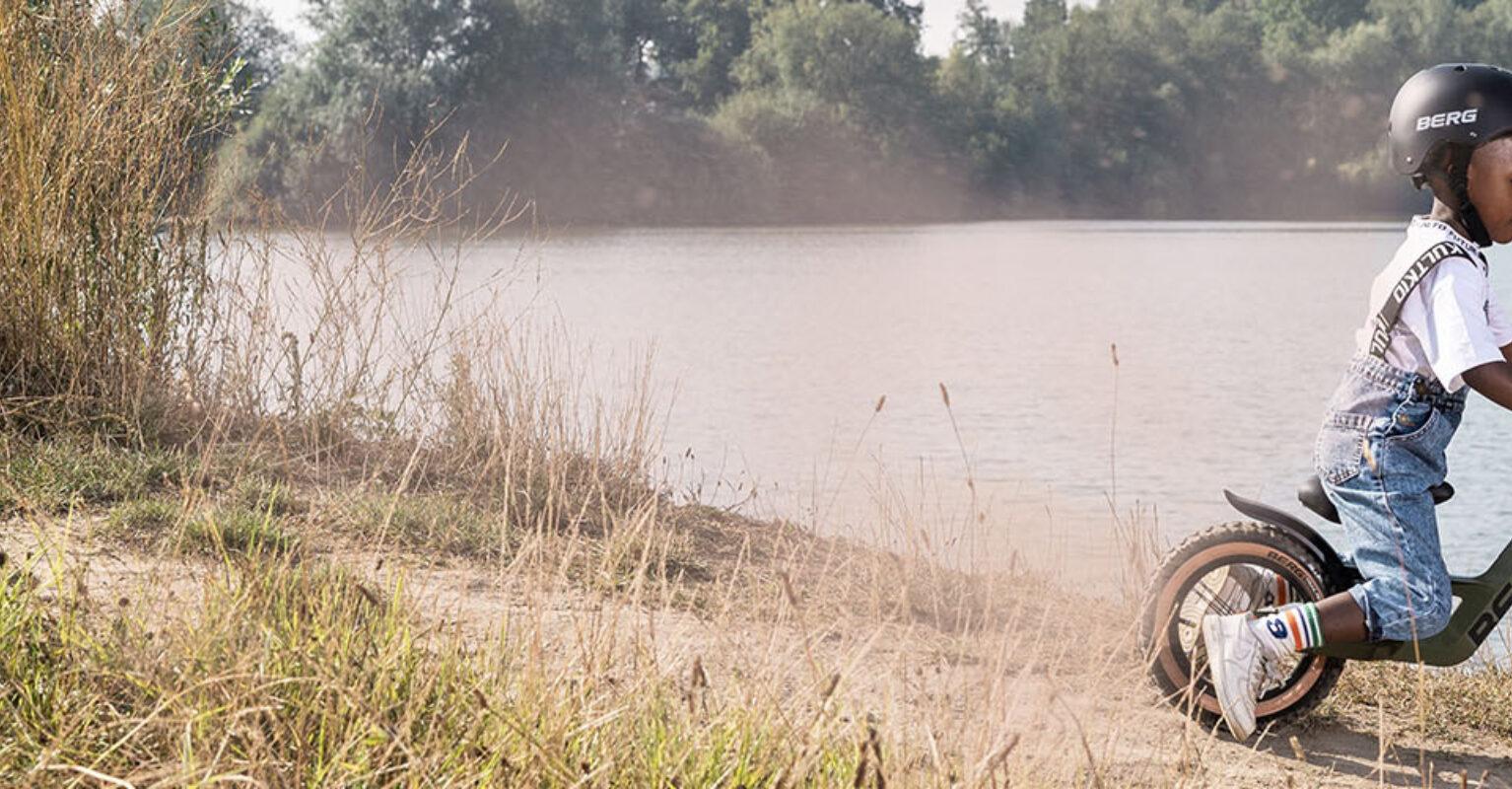 Banner with a child riding a BERG Biky Cross balance bike along the water on a sandy path