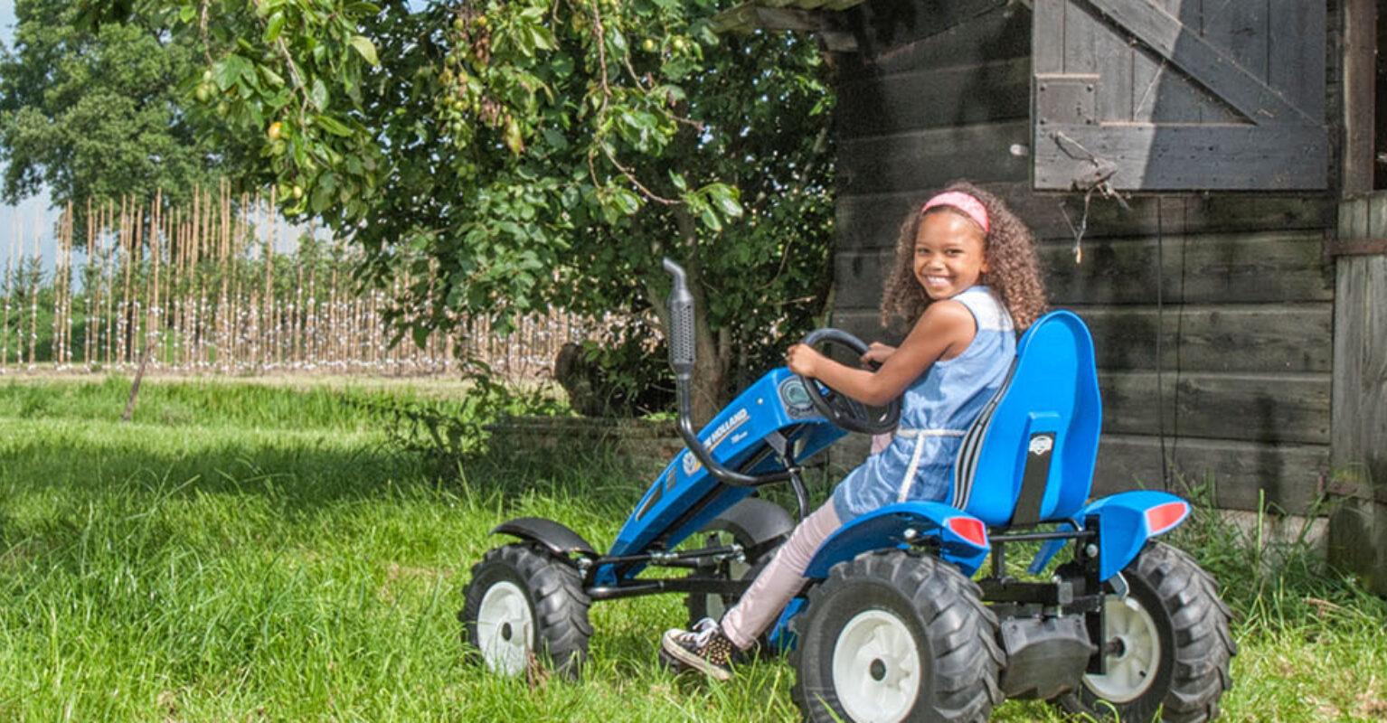 Child driving a BERG New Holland pedal go-kart near a wooden barn while another child watches, banner