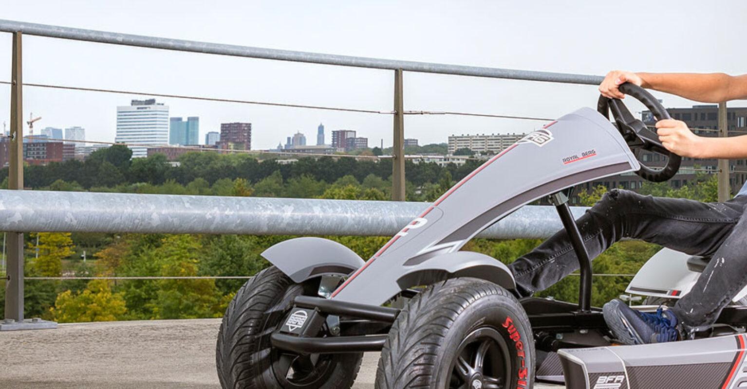 Garçon conduisant un kart à pédales BERG Race XL sur un pont surélevé avec vue sur la ville, bannière